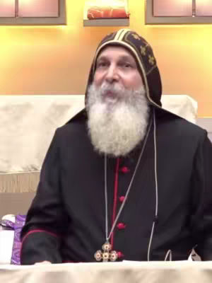 The image shows a man in religious attire, likely a bishop, sitting indoors. He has a white beard and a serious expression. He is wearing a black robe with a red collar and a black hood decorated with gold patterns.