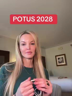 A woman with long blonde hair looks directly at the camera. Above her head is a red banner that says 'POTUS 2028.' She is holding a white object. The background is a room with a bed and a framed picture.