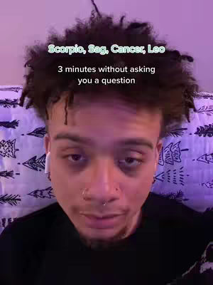 A person of color is centered in the frame, their head filling most of the space. They have short, dark, curly hair and are wearing a black shirt. White earbuds are in their ears, and a nose ring is visible. The person is positioned against a backdrop of a patterned comforter, with text overlaid at the top.