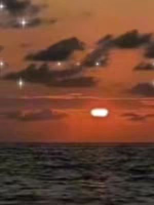 The image captures a sunset over the ocean. The sky transitions from a deep orange near the horizon to a lighter orange at the top, with some dark, cloud-like formations scattered throughout. The sun itself is a bright, horizontal oval shape, slightly obscured by the cloud cover. Below, the sea reflects the warm tones of the sky. The overall composition is serene and atmospheric.