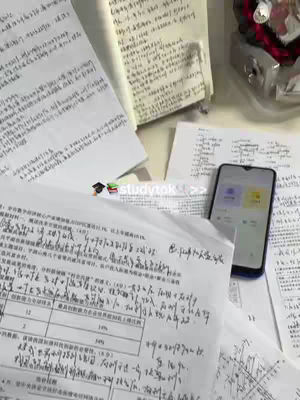 The image features a cluttered desk with various study materials, including scattered sheets of paper covered in handwritten notes and a partly visible open notebook. A smartphone displaying an app screen sits among the papers. The composition is top-down, highlighting the study environment, with muted lighting and a neutral color palette dominated by white and beige tones.