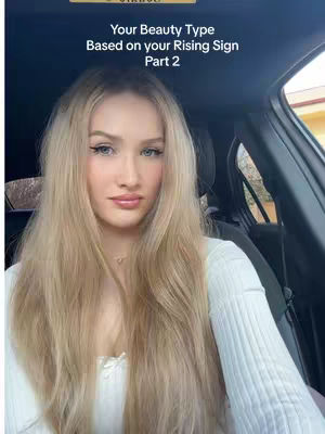 A woman with long blonde hair is sitting in the driver's seat of a car and looking directly at the camera. The background is blurred, showing the interior of the car.