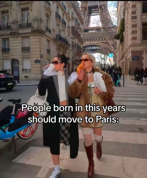 Two women stand in front of Parisian architecture. One is eating a pastry. The Eiffel Tower is visible in the background.
