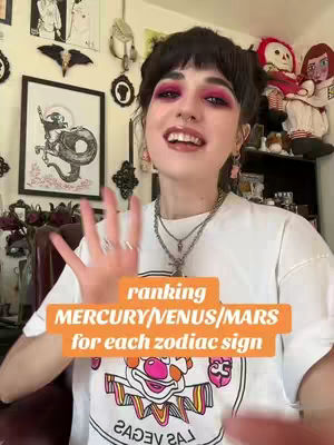 A woman with dark hair and bright makeup, is positioned in front of a wall filled with decorations. She is wearing a white t-shirt with a colorful design. An orange rectangle containing the text "ranking MERCURY/VENUS/MARS for each zodiac sign" is overlaid on the image.