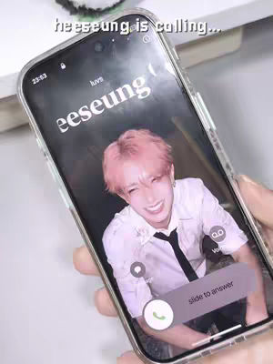 A phone held at an angle with the screen displaying a call from a person with pink hair and a white shirt, overlaid with a text hook. The background includes a blurred hand and white surface.