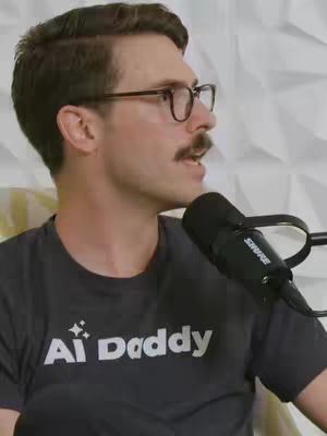 A man with brown hair, glasses, and a mustache speaking into a microphone. He is wearing a dark gray t-shirt with white text that reads 'Ai Daddy'. The background is a white, textured pattern.
