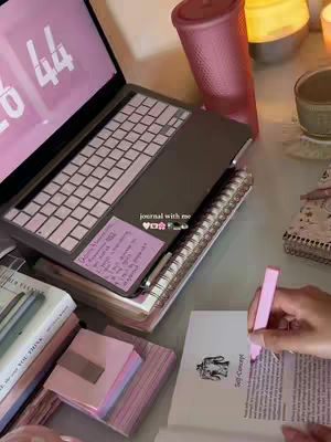 A desk setup with a laptop, notebook, books, and a pink cup. A hand is shown, holding a pink highlighter, highlighting words in a book with an image of an anatomically-correct man on a pedestal. The laptop screen displays what looks like a pink clock, other study materials and accessories are arranged in an organized display.