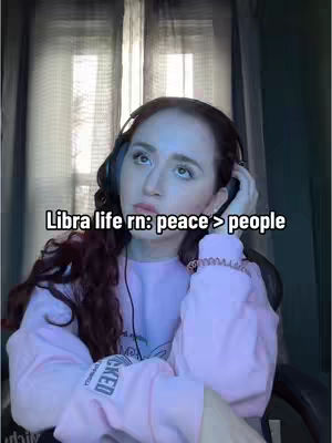 A woman with long, dark hair wearing a pink sweater and headphones looks upwards with a thoughtful expression. Text overlayed on the image reads: \"Libra life rn: peace > people\". The background shows a window, and the lighting suggests an indoor setting.