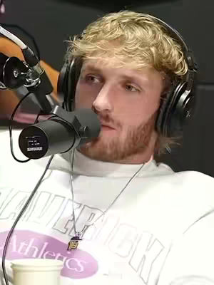 A close-up shot of a man in a studio setting wearing headphones and holding a microphone. He has blonde hair, a beard, and is wearing a white t-shirt. A necklace hangs around his neck.