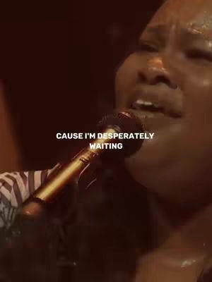 The image captures a close-up of a person singing into a microphone, bathed in warm, dim lighting. The person, seen from the chest up, has dark skin and is wearing a garment with a striped pattern on the upper left. They are mid-song, with their mouth open and eyes closed. The microphone is positioned close to the mouth, and the background is a blurred, dark hue, focusing attention on the singer's face. Text overlayed at the bottom states, 'CAUSE I'M DESPERATELY WAITING' in white font.