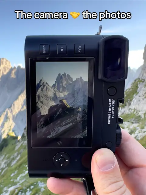 A close-up shot of a black Leica camera held against a backdrop of a mountain landscape on a clear day. The camera's screen displays a picture of the mountains, highlighting the scenic view. The camera is held by a person's hand, which is partially visible. The text overlay at the top reads 'The camera 🤝 the photos'.