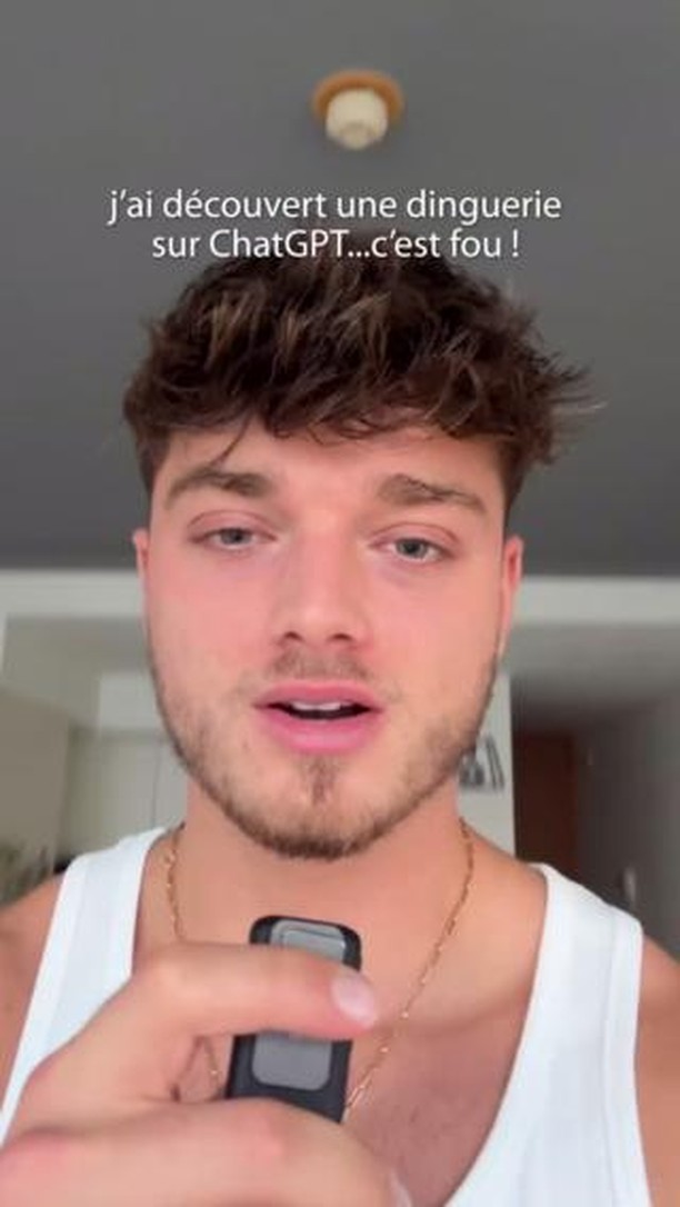 A selfie-style image featuring a young man with curly brown hair, fair skin, and a slight beard. He wears a white tank top and a gold chain. He is holding a small, black device in his right hand, and his expression appears to be one of surprise or excitement, with his mouth slightly open. The background is blurred, suggesting a room with a ceiling light. The text overlay reads, 'j'ai découvert une dinguerie sur ChatGPT...c'est fou !' 