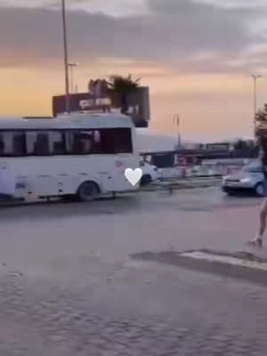 A woman walking on a wet street near a bus and other vehicles. The sky has shades of orange and pink.