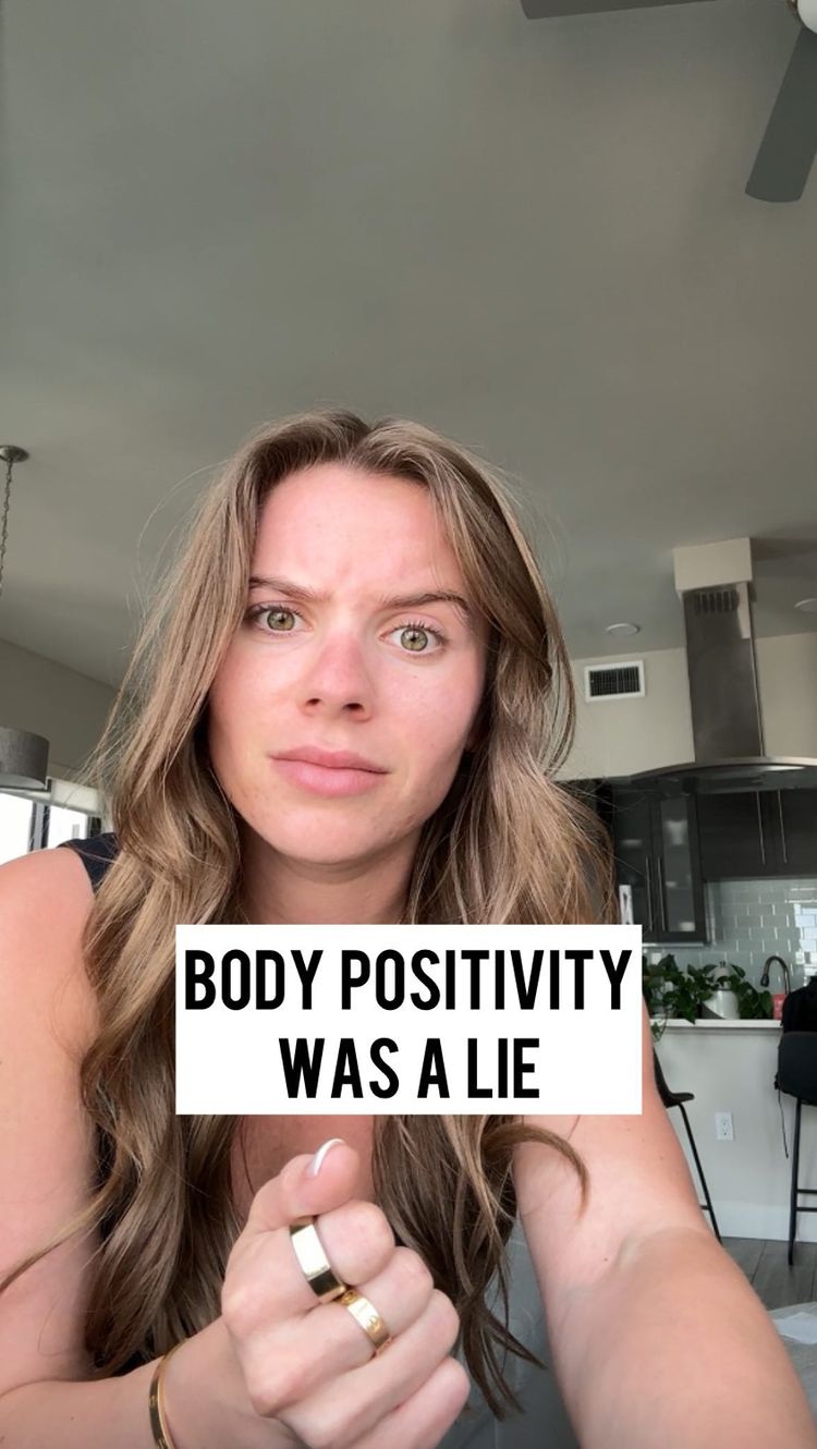 A woman with long blonde hair is looking directly at the camera with a pensive expression. A white rectangle with black text reading "BODY POSITIVITY WAS A LIE" is overlaid in the center of the shot. She is holding up a hand with several rings on her fingers and is positioned in a home setting, with a blurred-out kitchen visible behind her.