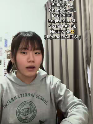 A young woman with dark hair in pigtails is centered in front of a gray wall, wearing a gray hoodie with the words 'INTERNATIONAL SCHOOL' on it. Text is superimposed on the right side of the frame, and the overall lighting is natural.