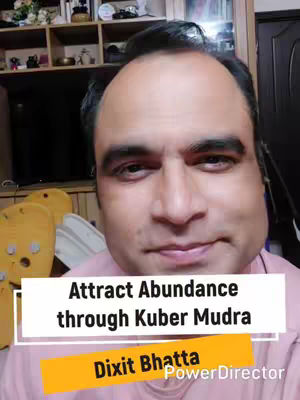 A man with a fair complexion, wearing a light pink shirt, is centered in the frame. The background features a room with wooden shelves displaying various items and a television. Overlayed across the image are text boxes with the words 'Attract Abundance through Kuber Mudra' and the creator's name, 'Dixit Bhatta', along with the 'PowerDirector' branding. The overall composition is a medium shot with the focus on the man's face.