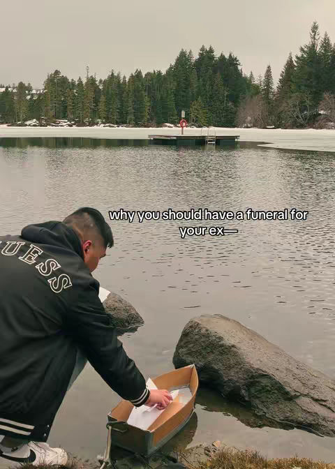 A man kneels by a body of water, placing a small boat with papers inside. The background includes a lake, snow, trees, and a dock. The color palette is muted, with grays, blues, and greens dominating the scene.