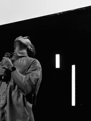 A black and white image of a man with his face tilted upwards, appearing to sing or speak into a microphone. The lighting suggests a stage setting. Behind him is a dark backdrop with white vertical rectangular lights. The composition is focused on the man.