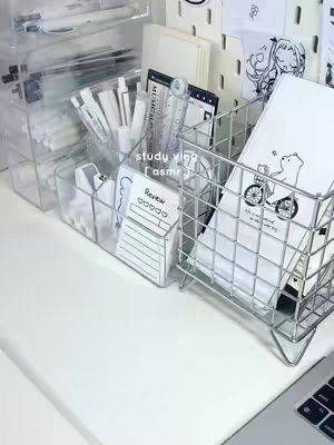 A neatly organized desk with various stationery items like pens, markers, and notebooks. There are clear acrylic containers and a wire basket to hold these items. The color scheme is predominantly white with touches of clear and gray, giving a clean and minimalist aesthetic.