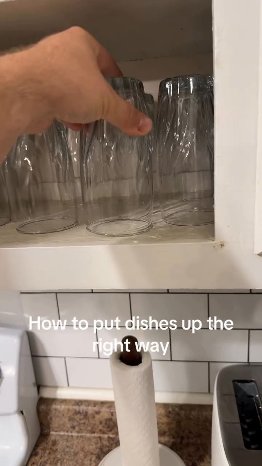 A hand is holding an upside down glass above a shelf in a kitchen cabinet. Below the shelf are white tiles. In the foreground is a roll of paper towels on a holder.