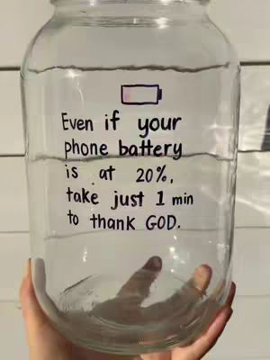 A hand holds a clear glass jar filled with water. Handwritten text on the jar reads, 'Even if your phone battery is at 20%, take just 1 min to thank GOD.' A small battery icon showing low charge is drawn near the top.