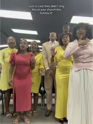 A group of people, likely a choir, standing indoors and singing. The people are dressed in formal or semi-formal attire. The image has a mixed lighting with some areas appearing brighter than others.