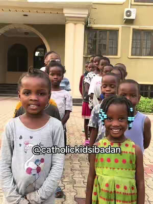 A group of children, mostly of African descent, are standing in a line in front of a building with a light-colored facade. The children are all looking towards the camera. The child in the front on the left has a gray shirt with a design, while the child on the right has a green dress with red dots. There is visible text overlay.