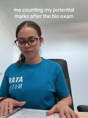 A young woman with glasses and a blue shirt is seated, looking down at a notebook with a pen. The background is a room with white walls and a window, and a text overlay indicates the topic is about the biology exam.