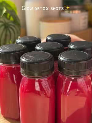 Close-up shot of several small, clear bottles filled with a vibrant pink-red liquid. The bottles have black caps. The bottles are arranged in a close cluster, taking up most of the frame. In the background, a blurred view of additional bottles and a green plant is visible, suggesting a kitchen or food preparation setting. The image is well-lit and focuses on the color and texture of the liquid.