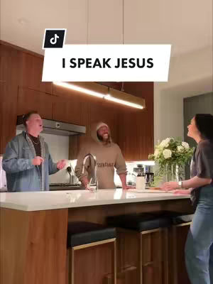 A vertical video shot in a well-lit kitchen. Three people are present: a person on the left dressed in a jean jacket, a person in the middle wearing a hoodie, and a person on the right in a casual outfit, all interacting near a kitchen island. The background includes kitchen cabinets, a stovetop with a hood, a countertop and a vase of flowers.