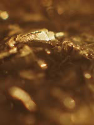 An extreme close-up shot of a surface that appears to be crystalline or mineral-like, with a warm, golden-brown color palette. The focus is soft, with blurred areas showcasing some bright, glinting highlights and bokeh effects adding depth and suggesting a play of light. The texture is rough and uneven, and the overall image gives a sense of depth or complexity.