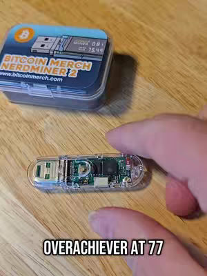 The image shows a close-up shot of a wooden surface with a clear plastic case and a small USB drive-like device, and a pair of hands. The top right corner features a blue case with the text 'Bitcoin Merch Nerdminer 2.' The device held is detailed with green and black electronic component, held by a person's fingers. At the bottom, 'OVERACHIEVER AT 77' is displayed in white text.