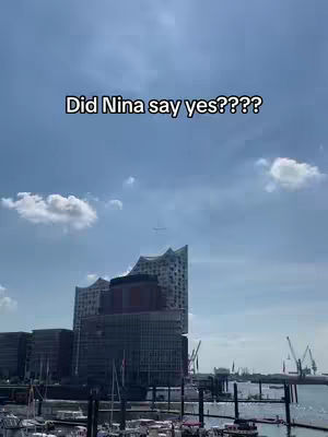A cityscape with a large modern building dominating the center, set against a blue sky with scattered clouds. The building has a unique architectural design, with a wavy roof. The sky is bright, suggesting a daytime setting. In the foreground, there are structures and water.