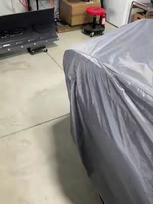 The image shows a garage with a car covered in a gray cover, the room is lit with natural light.