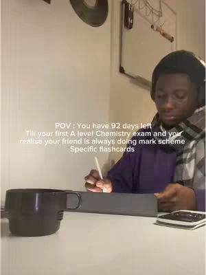 A young man with dark hair wearing a purple shirt and a scarf sits at a desk, focused on studying. There's a cup, a notebook, and a tablet on the desk.