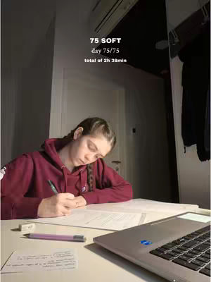 A young woman with braided hair sits at a desk, writing on paper. Her face is angled down, focused on the work. A laptop and other study materials are on the desk. Text overlays at the top indicate this is the 75 SOFT challenge, and also includes day and time information.