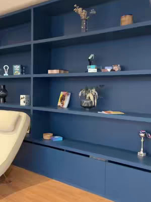 The image shows a well-organized, stylish interior, featuring a deep blue built-in bookshelf unit. The unit fills most of the frame, composed of shelves and cabinets. Various decorative items fill the shelves, adding visual interest. The room is lit and has wooden flooring.