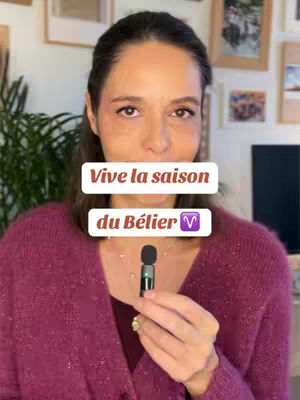 A woman with brown hair and a magenta sweater is holding a microphone in front of a blurred background with framed pictures, with two text bars on her face that say "Vive la saison" and "du Bélier ♈".