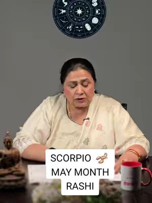 A woman sits at a table with a coffee mug, looking directly at the camera. Behind her, a zodiac wheel hangs on the wall. The image has a simple, direct composition with good lighting.