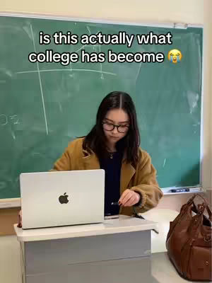 A woman with glasses sits at a school desk with a laptop open. Behind her is a chalkboard and a brown purse. The background is a classroom interior.