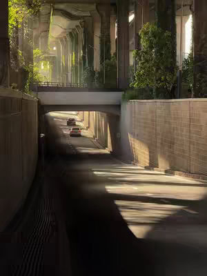 An architectural image showcasing an underground road or tunnel. A road is visible, leading toward the vanishing point.  The road is lined and divided into lanes. The tunnel or road is framed by towering pillars or walls, constructed from a light-colored material as though cast in concrete. Vegetation, likely plants or foliage, grows in patches along the sides and on top of the tunnel structure. Sunlight casts patterns of light and shadow. The light creates strong contrasts and adds depth to the shot, enhancing the architectural elements and the composition.