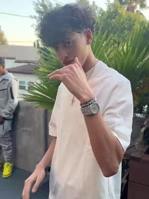A young man with dark curly hair is positioned in front of some green foliage. He's wearing a white t-shirt and has a watch and bracelet on his wrist. He's holding up his hand in a 'call me' gesture. Another person is partially visible in the background. 