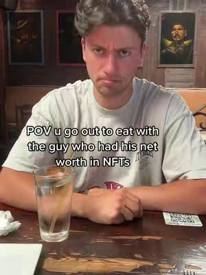 A man sits at a table in a restaurant, looking at the camera with a somewhat annoyed expression. In front of him is a glass of water and a QR code. The background includes artwork and a booth.