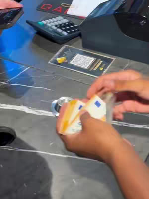 The cover image is a first-person perspective shot. The main focus is on the hands holding a stack of euro banknotes. The hands are dark-skinned, holding approximately 50 Euro bills. The background features a marble-like countertop, a calculator, and parts of a cash register device.