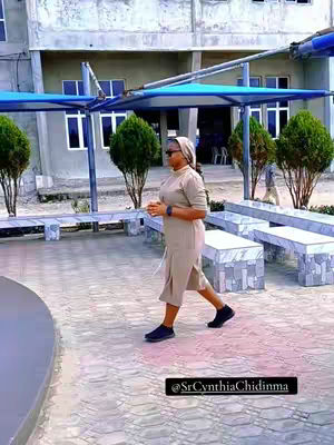 A woman in a beige dress and head covering is walking along a paved area. She is wearing sunglasses and carrying her hands in front of her. The background includes windows, trees, benches, and a shaded structure overhead. The photo is well lit and has a calm, neutral color palette.