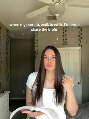 A young woman stands in a bedroom, looking at the camera and holding a book. Text overlayed on the image reads 'when my parents walk in while I'm tryna share the Bible'. The composition is a medium, shot with the top of a ceiling fan visible in the background, and the head and shoulders of the woman as the focal point.