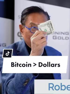 A middle-aged man wearing glasses holds a one-dollar bill up to his eye, obscuring part of his face. He is dressed in a blue blazer, and the background is slightly blurred, with text that reads 'MINING GOLD'. A white banner with the words 'Bitcoin > Dollars' is overlaid on the bottom portion of the image.