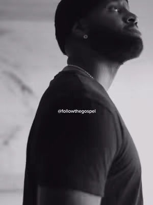 A grayscale image features a man from the chest up, seen in a three-quarter profile. He has a short haircut, a closely trimmed beard, and wears a dark-colored t-shirt. A silver chain necklace is visible. The background is plain and out of focus.