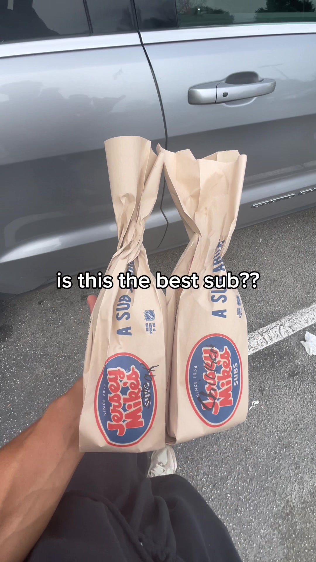 A photo shows a close-up of two Jersey Mike's sub sandwiches in their brown paper bags, held by a person in front of a silver car. The bags have the Jersey Mike's logo visible, with the phrase "is this the best sub??" imposed in white letters overlaid.