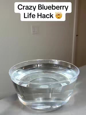 A clear plastic bowl filled with water sits on a white surface. Above the bowl, text in a white box reads "Crazy Blueberry Life Hack" with a small emoji of a cooked pot or pan.